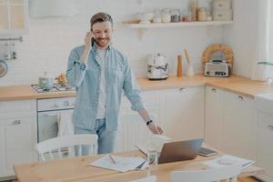 Schöner lächelnder bärtiger Mann, der auf dem Handy in der Küche spricht, während er aus der Ferne arbeitet foto