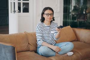 frau mit brille, die fernsehkanäle wechselt, fernsieht, sich ausruht, zu hause auf dem sofa sitzt foto