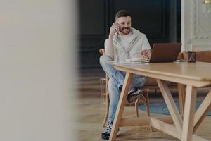 Junger lachender Freiberufler, der mit dem Handy spricht, während er zu Hause an seinem Arbeitsplatz sitzt und remote am Laptop arbeitet foto