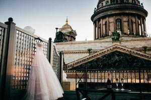 schönes weißes Hochzeitskleid auf Kleiderbügeln. Champagner und zwei leere Gläser auf dem Tisch. Hochzeitsvorbereitung. Hochzeitszubehör vor Kathedralenhintergrund. elegantes Kleid der Braut. Hochzeitskonzept foto