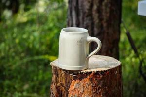 Große weiße Keramik-Biertasse mit Griff, die auf geschnittenem Baumstumpf im Waldgras steht foto