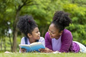 Die afroamerikanische Mutter bringt ihrer kleinen Tochter das Lesen bei, während sie sich hinlegt, nachdem sie ein Sommerpicknick im öffentlichen Park für Bildung und Glückskonzept hatte foto