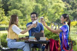 Eine Gruppe von College-Studenten trifft sich und arbeitet im Sommer draußen im Garten des Universitätscampus an Abschlussarbeiten und Projekten, während sie mit heißem Kaffee zum Mitnehmen jubelt foto