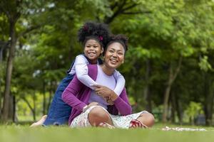 afroamerikanische mutter spielt huckepackreiten und umarmt sich mit ihrer kleinen tochter, während sie ein sommerpicknick im öffentlichen park für das wohlbefinden und glückskonzept hat foto