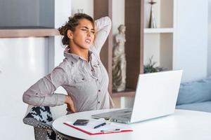 porträt einer jungen gestressten frau, die zu hause am schreibtisch vor dem laptop sitzt, den schmerzenden hals mit schmerzendem ausdruck berührt und nach der arbeit am pc unter nackenschmerzen leidet foto