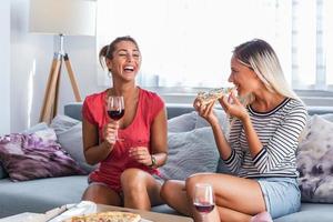 glückliche junge freundinnen, die zu hause pizza mit wein auf dem sofa essen. Fröhliche, lächelnde junge Freundinnen, die Pizza essen und zu Hause eine Flasche Wein öffnen foto