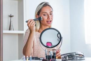 Schönheitsfrau, die Make-up anwendet. Schönes Mädchen, das in den Spiegel schaut und Kosmetik mit einem großen Pinsel aufträgt. Mädchen wird auf den Wangenknochen rot. Puder, Rouge foto