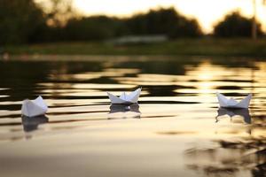 Drei Papierboote, die bei einem wunderschönen Sonnenuntergang auf den Wellen im Wasser schwimmen. Abends selbstgemachte Origami-Boote auf der Wasseroberfläche. Nahansicht. foto