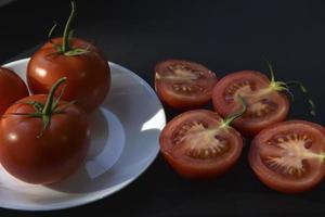 schöne früchte einer ganzen und geschnittenen tomate auf einer weißen plattenseitenansicht. Köstliches Tomaten-Stillleben in den Strahlen der Abendsonne auf schwarzem Hintergrund. foto