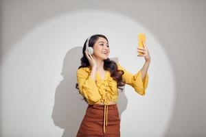 Porträt eines jungen, fröhlichen Mädchens mit weißem Kopfhörer macht Selfie auf dem Smartphone, das über weißem Hintergrund isoliert ist foto