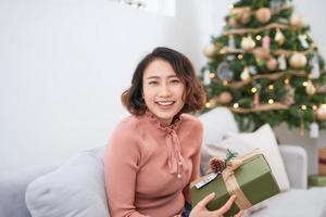 lächelnde frau mit geschenkbox vor dem hintergrund des weihnachtsbaums. glückliche junge frau, die weihnachten feiert foto