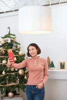 lustiges asiatisches mädchen, das selfie-bilder auf smartphone-kamera zu hause nahe weihnachtsbaum macht foto