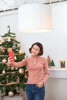 urlaub, winter und personenkonzept - glückliche junge frau, die zu hause selfie in der nähe des weihnachtsbaums macht foto