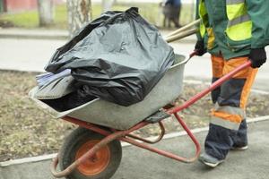 Arbeiter trägt Müllwagen. Arbeitskleidung in hellen Farben. Wagen mit Taschen. foto