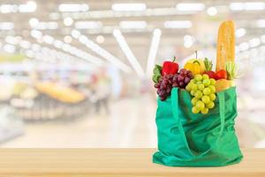 frisches obst und gemüse in wiederverwendbarer grüner einkaufstasche auf holztisch mit supermarkt-lebensmittelgeschäft unscharfer defokussierter hintergrund foto