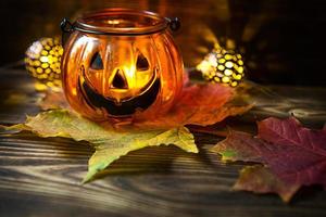 Lampenkürbis mit Augen und Mund aus Glas und natürlichem orangefarbenem Kürbis auf einem Holztisch mit gelben und roten Ahornblättern. Halloween, warme Herbstatmosphäre. runde Girlande, Kerze in einem Leuchter. foto