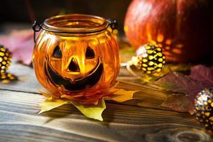 Lampenkürbis mit Augen und Mund aus Glas und natürlichem orangefarbenem Kürbis auf einem Holztisch mit gelben und roten Ahornblättern. Halloween, warme Herbstatmosphäre. runde Girlande, Kerze in einem Leuchter. foto