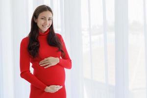 Eine schöne, kühle, schwangere Frau stand mit ihren Händen auf ihrem Bauch und berührte das ungeborene Kind zärtlich, wertschätzend und glücklich mit einem lächelnden Gesicht foto