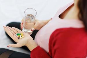 schwangere Frau, die ein Glas Wasser hält und die Anzahl der Pillen zählt, die sie in der Hand hält, um ihren Körper zu ernähren. foto