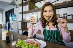 Matue Erwachsene asiatische Hausfrau in Schürze, die am Tisch in der Küche sitzt und ein Glas Milch hält und bereit ist, Frühstück zu essen. glückliche süße frau, die den neuen tag morgens mit leckerem essen beginnt foto