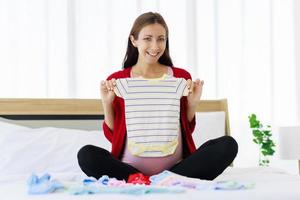 Eine große schwangere Frau bereitet glücklich Babykleidung auf ihrem Bauch vor. foto