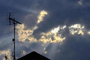 Sonnenstrahlen ragen am späten Nachmittag durch dunkle Wolken. Dach- und Fernsehantenne im Vordergrund. foto
