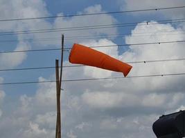 orangefarbener Windsack am blauen Himmel mit ein paar flauschigen Wolken, die meistens gerade heraus wehen. Um den Windsack herum befindet sich ein elektrisches Kabel. foto