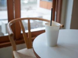 Eismilchkaffee in weißem, leerem Pappbecher auf dem Tisch in der Nähe des Glasfensters in einem minimalen Café im japanischen Stil foto