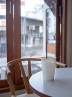 Eismilchkaffee in weißem, leerem Pappbecher auf dem Tisch in der Nähe des Glasfensters in einem minimalen Café im japanischen Stil foto