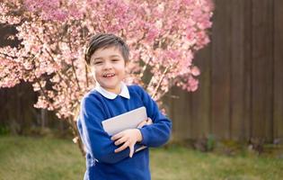 porträt glückliches kind mit tablet-pc, das morgens auf den schulbus wartet, positiver kleiner junge mit schuluniform, der in die kamera schaut, während er im garten steht. bildung und zurück zum schulkonzept foto