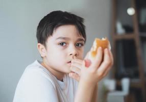 Hungriger kleiner Junge, der hausgemachte Specksandwiches mit gemischtem Gemüse isst, gesundes Kind, das zu Hause frühstückt, Kind, das Fingernägel beißt und mit denkendem Gesicht herausschaut foto