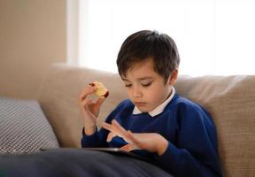 glücklicher Schuljunge, der roten Apfel isst, während er online mit einem Freund auf dem Tablet spielt, Kind, das dem Lehrer Hausaufgaben über das Internet schickt, positives Kind, das auf dem Sofa sitzt und sich allein im Wohnzimmer entspannt foto