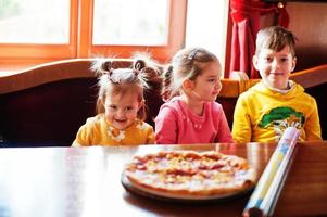 Kinder an Geburtstagen sitzen am Tisch und essen Pizza. foto