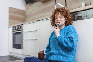 süße ingwerjunge frau in modernen, stilvollen kleidern, die leckeren schokoladenaufstrich mit süßem lächeln in der küche genießen. junge Frau, die Schokolade aus einem Glas isst, während sie auf dem Küchenboden sitzt. foto