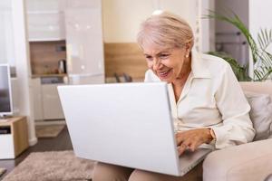 ältere dame, die mit laptop arbeitet. Porträt einer schönen älteren Frau, die drinnen mit einem Laptop arbeitet. ältere frau, die zu hause laptop verwendet und lacht foto