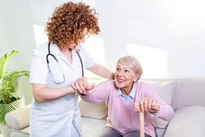 weibliche Betreuerin hilft Seniorin beim Aufstehen von der Couch im Wohnzimmer. lächelnde Krankenschwester hilft Seniorin beim Aufstehen. Fürsorgliche Krankenschwester unterstützt den Patienten beim Aufstehen vom Sofa foto