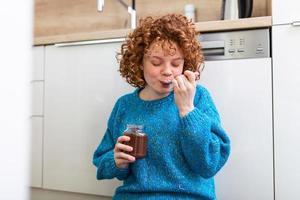 junge Frau, die Schokolade aus einem Glas isst, während sie auf dem hölzernen Küchenboden sitzt. süßes Ingwermädchen, das sich mit einem frechen Gesicht verwöhnt und Schokoladenaufstrich aus dem Glas mit einem Löffel isst und jeden Bissen genießt foto