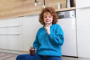 süße ingwerjunge frau in modernen, stilvollen kleidern, die leckeren schokoladenaufstrich mit süßem lächeln in der küche genießen. junge Frau, die Schokolade aus einem Glas isst, während sie auf dem Küchenboden sitzt. foto