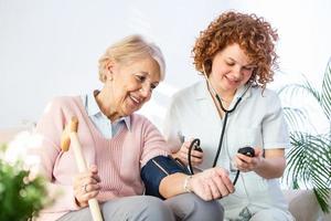 glückliche Seniorin, die ihren Blutdruck in einem Pflegeheim von ihrer Pflegekraft messen lässt. Glückliche Krankenschwester, die den Blutdruck einer älteren Frau im Wohnzimmer misst foto