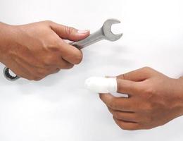 Nahaufnahme Hand asiatischer männlicher Techniker, der einen Werkzeugschlüssel hält, ist in Gefahr. und Verletzungen der Finger durch die Arbeit in Industrieanlagen mit Erster Hilfe mit Verband im Krankenhaus auf weißem Hintergrund.
