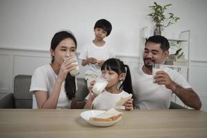 eine gesunde asiatische thailändische familie, kleine kinder und junge eltern trinken morgens frische weiße milch im glas und brotfreude zusammen an einem esstisch, wellness ernährung zuhause frühstücksmahlzeit lebensstil. foto