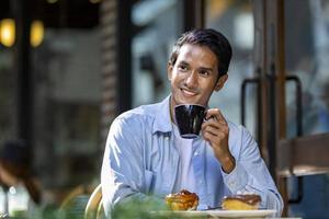 asiatischer mann, der einen heißen espressokaffee schlürft, während er vor dem café-bistro im europäischen stil sitzt und das langsame leben mit morgenstimmung auf dem stadtplatz mit süßem gebäck genießt foto