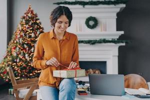 Lächelnde frohe italienische Frau, die rotes Band auf Weihnachtsgeschenk bindet, während sie zu Hause im Büro sitzt foto