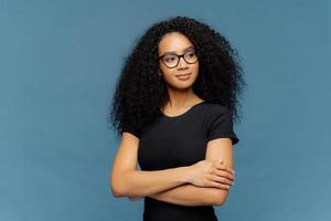 Foto einer nachdenklichen, zufriedenen Afro-Frau hält die Hände über der Brust gekreuzt, fokussiert beiseite, trägt eine transparente Brille, ein lässiges schwarzes T-Shirt, isoliert auf blauem Hintergrund. Konzept der menschlichen Gesichtsausdrücke