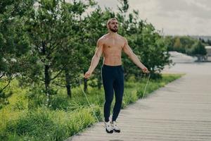 sportlicher mann mit dickem bart macht übungen mit springseil, hat muskulösen oberkörper, bleibt in guter körperlicher form, posiert im freien. Fitness und gesundes Konzept. männlicher läufer wärmt sich mit sportgeräten auf foto