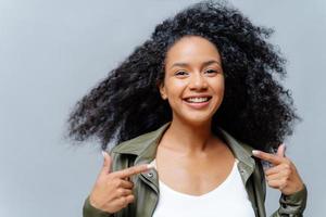 kopfschuss einer hübschen lächelnden frau mit afrofrisur, zeigt mit beiden zeigefingern auf sich selbst, fühlt sich stolz auf sich selbst, trägt lederhemd, isoliert über studiohintergrund. Schau mich an, mein neues Outfit foto