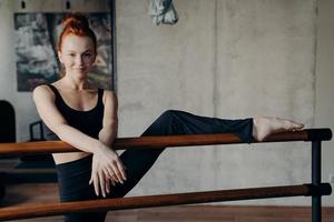 athletische lächelnde rothaarige frau, die sich auf der ballettstange ausdehnt foto