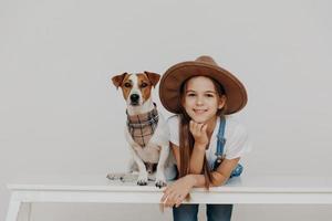 Zufriedenes hübsches Mädchen trägt einen Hut, lehnt sich an einen weißen Tisch, posiert in der Nähe eines Rassehundes, genießt die gemeinsame Freizeit, geht spazieren, ruht sich zu Hause aus. Kinder, Glück, Tiere, Lifestyle-Konzept foto