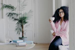 positive afroamerikanische geschäftsangestellte sitzt auf fensterbank, hält tasse kaffee, gekleidet in formelle kleidung, posiert in der nähe des desktops mit büchern, grüne zimmerpflanze im hintergrund, hat pause foto