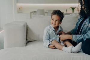 Fürsorgliche afroamerikanische Mutter verkleidet kleinen Sohn, während er im Wohnzimmer auf dem Sofa sitzt foto